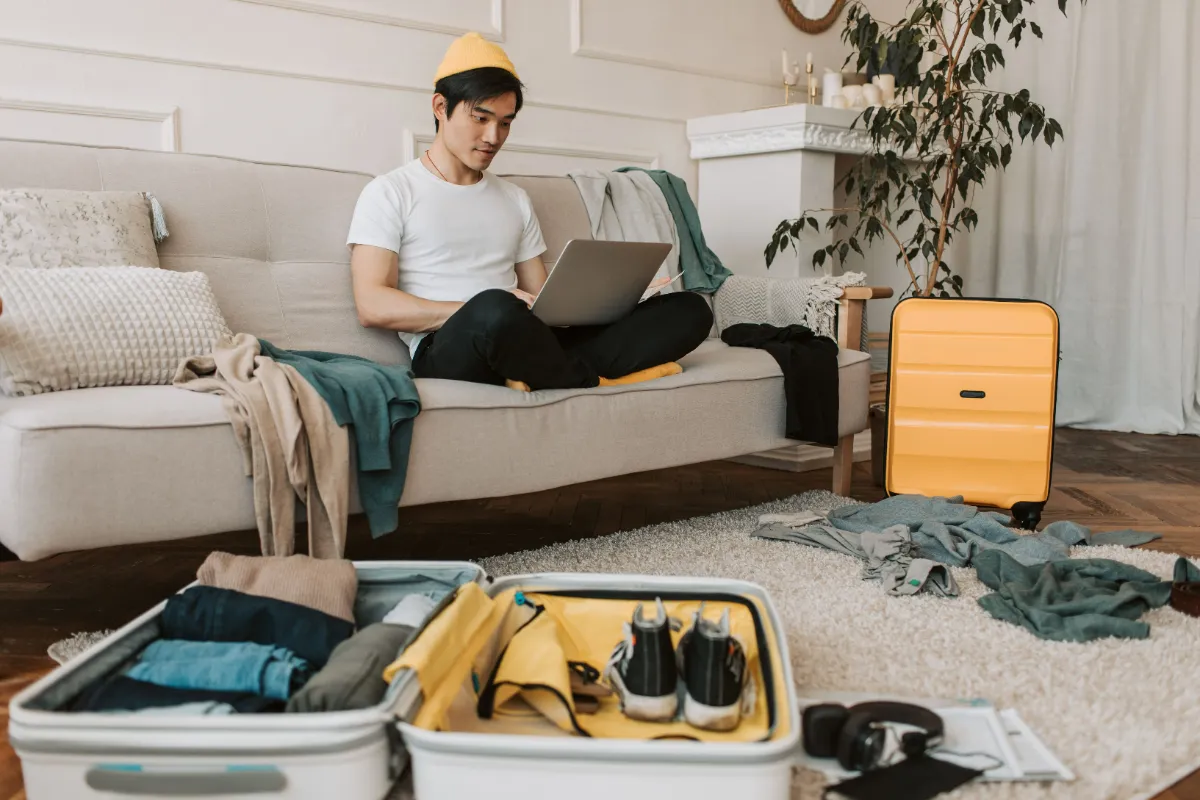A person sitting on a sofa with a laptop while an open suitcase and clothes are spread across the floor